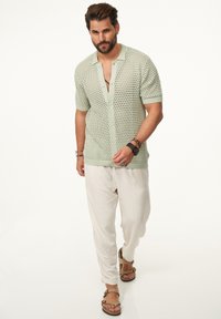 Hellgrüner, kurzärmeliger Strickcardigan mit Knopfverschluss und offenem Webmuster, kombiniert mit lockeren, off-white Leinenhosen und braunen Sandalen.