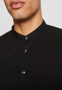 Czarna koszula z kołnierzykiem i listwą na guziki, z okrągłymi guzikami oraz gładką, matową fakturą. Widok z bliska na linię szyi.