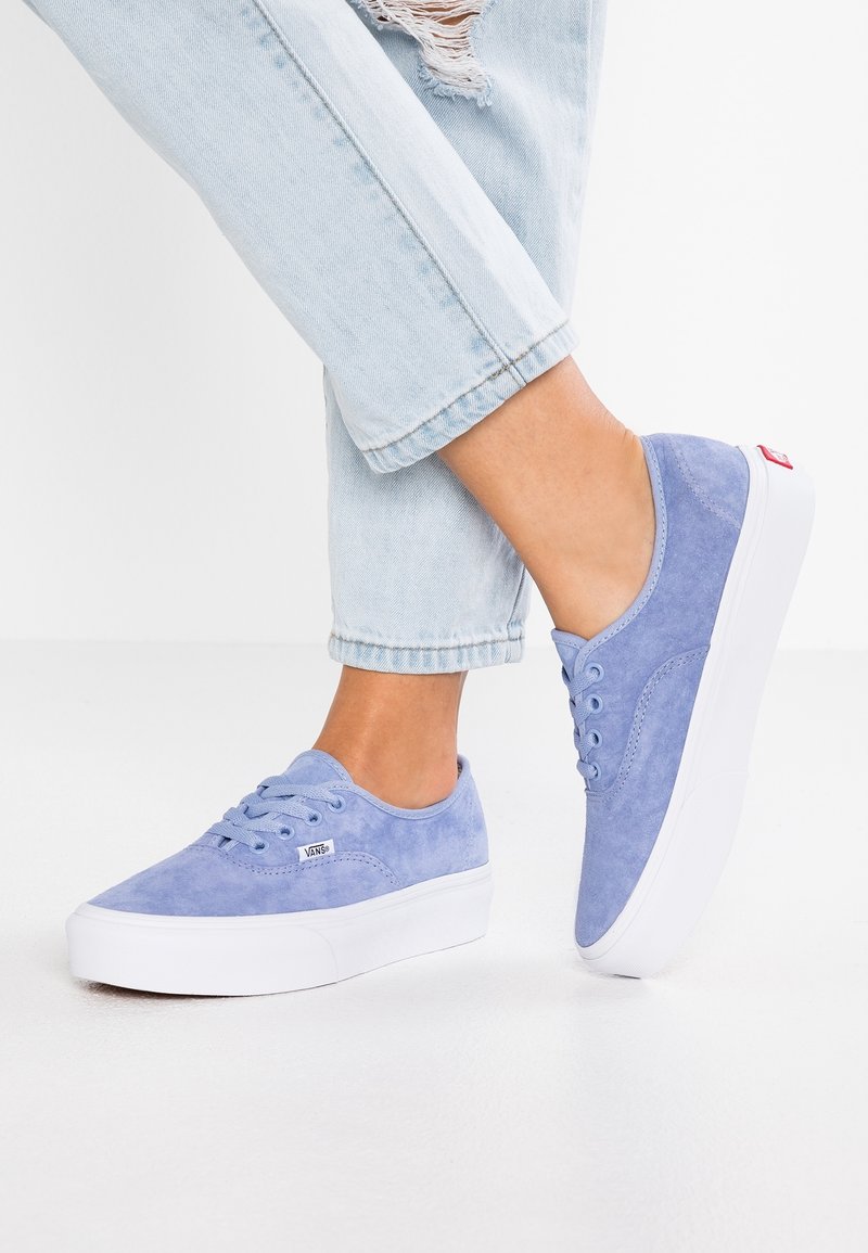 Personne portant des baskets Vans en daim bleu clair avec des semelles épaisses blanches et un jean bleu clair usé, debout sur une surface blanche.