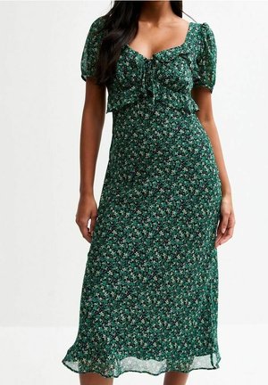 Vrouw draagt een groene midi-jurk met bloemenprint, opgeblazen mouwen en een gerimpelde, gestrikte taille, staande tegen een effen achtergrond.