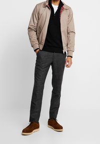 Giacca bomber beige con fodera a quadri rossa, maglione nero, pantaloni grigi con texture e scarpe in suede marrone con suola bianca.