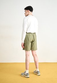 Pantalones cortos verdes con una cinturilla elástica, de una tela ligera, combinados con una camiseta blanca de manga larga y zapatillas grises con patrón.