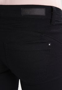 Jeans de mezclilla negra con una textura suave, que presentan un bolsillo trasero y una etiqueta de cuero con marca en la cintura. Detalle de costura simple.