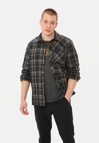 Camicia a quadri nera e grigia con dettagli gialli, con bottoni frontali, maniche arrotolate, indossata sopra una maglietta grigio scuro e pantaloni neri.