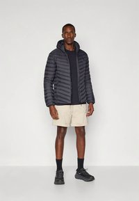 Veste matelassée noire à motifs horizontaux, fermeture éclair à l'avant et capuche attachée. Portée sur un T-shirt sombre et un short beige, complétée par des baskets noires.