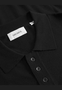 Zwarte gebreide poloshirt van Les Deux, met een opstaande kraag, drie zwarte knopen en een label met de merknaam en maat.
