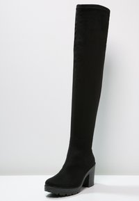 Bota negra hasta el muslo hecha de tejido suave, con un tacón grueso, punta redonda y suela texturizada con detalles de labrado.
