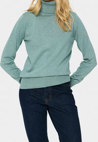 Pull col roulé vert sarcelle clair en tissu maille douce, avec manches longues et ourlet côtelé, associé à un jean en denim foncé.