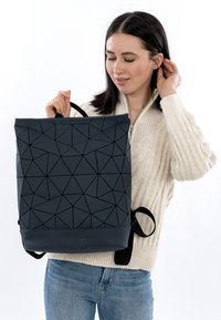 Svart ryggsäck med geometriskt mönster, texturerad yta, robusta handtag och justerbara remmar. Bärs av en person i en cremefärgad tröja.