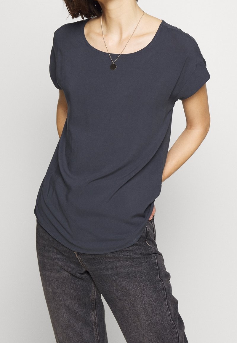 Personne portant une chemise à manches courtes bleu marine foncé avec un col rond et un jean noir, debout les mains derrière le dos.