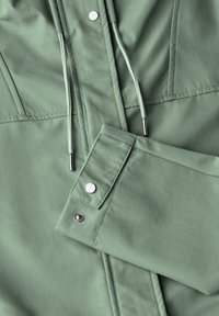 Veste verte à boutons, avec une texture lisse, featuring des cordons de serrage réglables, des fermetures à pression en métal, et un col et des manches structurés.