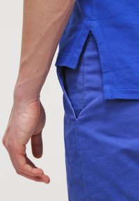 Blå kortärmad skjorta med en texturerad väv. Har sidoflikar och en ficka på vänster sida, i kombination med blå byxor.