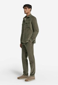 Olivgrön kordjacka i kord som har två bröstfickor, i kombination med matchande kordbyxor och beige sneakers. Slät textur och åtsittande design.