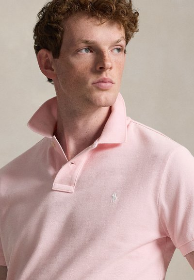 Νεαρός με σγουρά κόκκινα μαλλιά, φορώντας ανοιχτό ροζ πόλο μπλούζα με ανασηκωμένο γιακά, κοιτάζει στο πλάι με φόντο ουδέτερο.