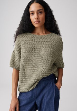 Femme aux longs cheveux bouclés portant un pull en tricot ample vert olive et un pantalon bleu foncé, debout avec une main dans la poche.