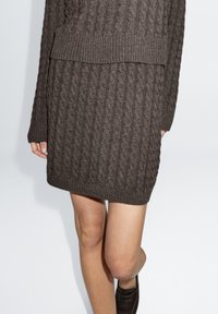 Gestricktes braunes Kleid mit Zopfmuster und geripptem Saum, mit langen Ärmeln und lockerem Schnitt. Material wirkt weich und strukturiert.