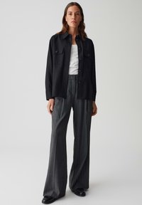 Camicia nera a maniche lunghe con due tasche sul petto, indossata sopra un top bianco. Pantaloni a gamba larga grigi a quadri e scarpe nere completano l'outfit.