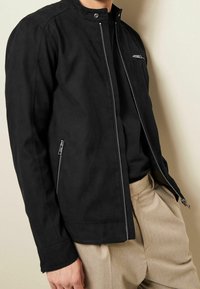 Schwarze Wildlederjacke mit hohem Kragen, seitlichen und Brust-Reißverschlusstaschen sowie silbernen Hardware-Akzenten. Getragen über einem schwarzen Oberteil und beigen Hosen.