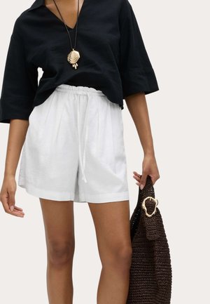 Frau trägt locker sitzendes schwarzes Oberteil, weiße Shorts mit Kordelzug, Muschelanhänger als Halskette, hält eine dunkle, gewebte Handtasche mit goldener Schnalle.