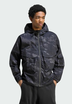Homme portant une veste à capuche camouflage noire et grise avec poches avant, fermeture éclair et poignets élastiques, mains dans les poches du pantalon.