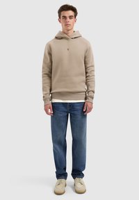 Jonge man in een beige hoodie, blauwe jeans en beige schoenen, staande tegen een effen lichtgrijze achtergrond.