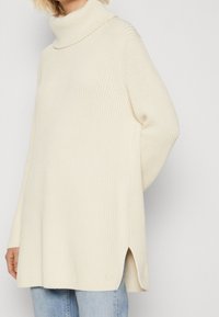 Person trägt einen cremefarbenen, gerippten Rollkragenpullover mit seitlichen Schlitzen, kombiniert mit hellblauen Jeans, vor einem schlichten Hintergrund.