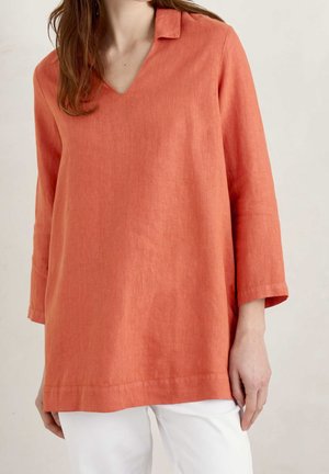 Kobieta ubrana w luźną, długorękawową, koralowo-pomarańczową bluzkę z dekoltem w serek i małym kołnierzykiem, zestawioną z białymi spodniami na jednolitym tle.