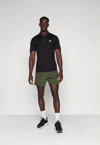 Svart kortermet poloshirt med glidelåshals og logo, kombinert med grønne shorts. Modellen har på seg svarte treningssko og hvite sokker.
