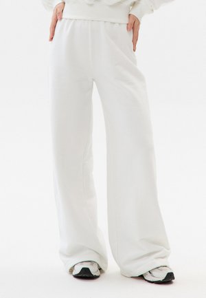 Osoba ubrana w luźne białe spodnie i białe trampki, stojąca z rękami opartymi na biodrach na białym tle.