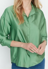 Satin grön skjorta med knäppning och krage samt långa ärmar. Slät textur med knappar fram och avslappnad passform.
