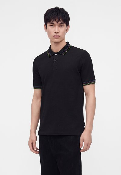 Jeune homme portant un polo noir à manches courtes avec un liseré rayé multicolore au col et aux poignets sur fond uni.