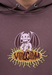 Sweat à capuche violet avec un motif brodé d'un chien cartoon aux ailes de chauve-souris au-dessus d'un logo circulaire portant l'inscription "VOLCOM" en marron et jaune.