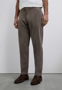 Pantalones de pana marrones con corte recto, que presentan pliegues, cierre de botón y textura de canalones verticales visibles. Llevados con zapatos oscuros.