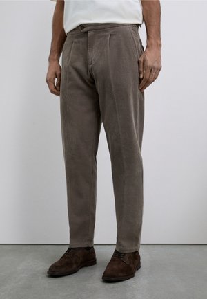 Pantalones de pana marrones con corte recto, que presentan pliegues, cierre de botón y textura de canalones verticales visibles. Llevados con zapatos oscuros.