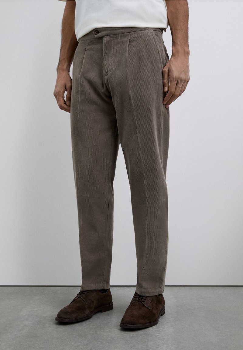 Pantalones de pana marrones con corte recto, que presentan pliegues, cierre de botón y textura de canalones verticales visibles. Llevados con zapatos oscuros.