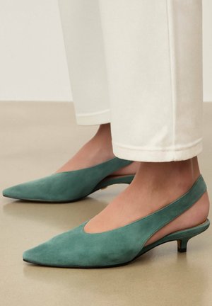 Tamsiai žalioline zomšine medžiaga padengti žemi kulniukai su siauru apsiuvu ir atvira kulno dalimi, dėvimi su baltais trumpais siaurais džinsais ant lygaus smėlio spalvos grindų.