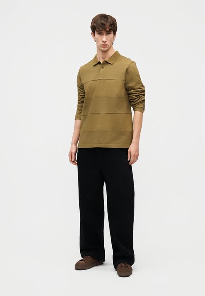 Νεαρός άνδρας στέκεται, φοράει πουκάμισο πόλο με μακριά μανίκια σε ελιά πράσινο χρώμα, μαύρο παντελόνι με φαρδιά πόδια και καφέ αθλητικά παπούτσια, με φόντο ένα απλό ανοιχτόχρωμο φόντο.