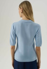 Žena s dlhými blond vlasmi, oblečená v svetlo modrom pletenom krátkom rukáve a tmavomodrých nohaviciach, otočená chrbtom k jednému svetlému pozadiu.