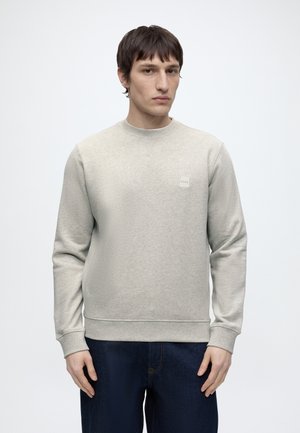 Fiatal férfi világosszürke pulóvert visel kis négyzet alakú logóval a mellkasán, sötétkék farmerral párosítva, egy sima háttér előtt áll.