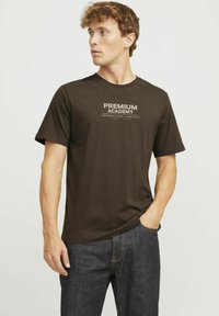 Camiseta de algodón marrón de manga corta, con un diseño de texto impreso "ACADEMIA PREMIUM" en la parte delantera. Combinada con jeans de mezclilla oscura.