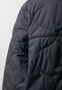Close-up of dark quilted fabric with curved stitching patterns, showing puffed texture and seam details on a garment.