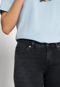 Camisa de algodão azul clara com gola redonda, combinada com jeans escuros de denim preto, apresentando um design de cinco bolsos e detalhes de metal visíveis na cintura.