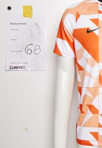Camisa deportiva Nike de color naranja y blanco con un diseño estampado exhibida en un maniquí, acompañada de una nota que muestra las medidas de hombros y longitud.