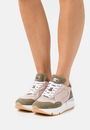 Jambes portant des baskets à lacets roses et olive avec des détails en mesh et en daim sur fond blanc.