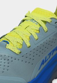 Κοντινό πλάνο αθλητικού παπουτσιού με κίτρινα κορδόνια, φωτεινή γκρι επιφάνεια με τρυπήματα και μπλε σόλα με το λογότυπο "Altra".