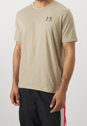 Homme portant un t-shirt beige Under Armour à manches courtes et un pantalon de sport noir avec des bandes latérales rouges et blanches, debout devant un fond uni.