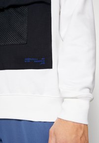 El sudadera blanca presenta un parche negro con detalles de malla, que muestra texto azul. Los puños y el dobladillo acanalados brindan estructura al diseño.