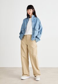 Giacca di jeans con bottoni a scatto, colore azzurro chiaro, indossata sopra una maglietta bianca. Pantaloni beige, di vestibilità ampia e sneakers bianche con plateau completano il look.