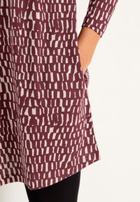 Nahaufnahme einer Hand in der Tasche eines weinrot-weißen gemusterten Langarmkleides, mit darunter sichtbaren schwarzen Leggings.