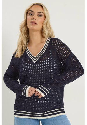 Lyshåret kvinde iført en marineblå, åbenstrikket sweater med hvide stribede manchetter, kant og V-hals, kombineret med lyseblå jeans.